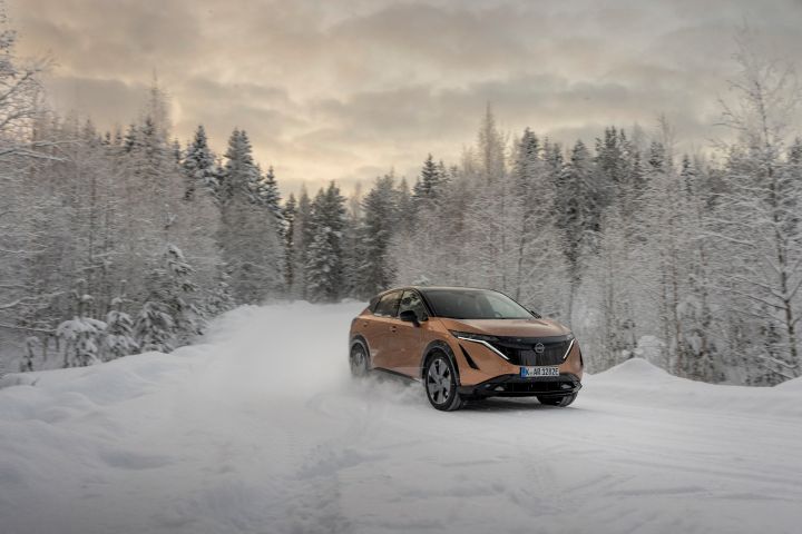 Elektroauto Nissan Ariya e‑4ORCE in Akatsuki Copper mit Dach und Außenspiegel in Pearl Black fährt auf schneebedeckter Straße mit verschneiten Bäumen auf beiden Seiten, 3/4 Front