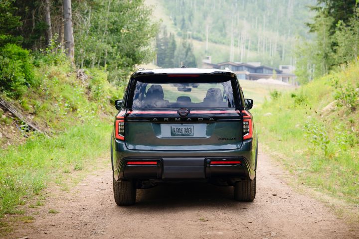 Rear view of a gray 2025 Nissan Armada parked on dirt path in the woods, cabins can be seen in the distance.