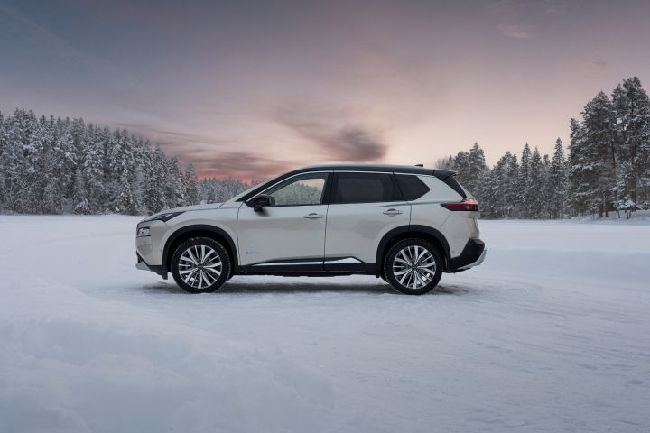 Silberner Nissan X‑Trail e‑Power e‑4ORCE steht auf schneebedecktem Eissee mit verschneiten Bäumen im Hintergrund, Seitenansicht