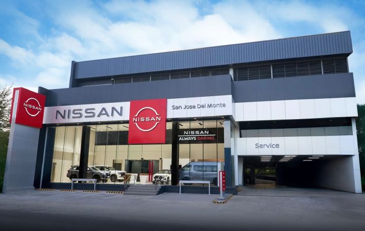 Front view of the Nissan San Jose Del Monte dealership featuring a modern gray-and-white facade, large glass showroom displaying vehicles, red Nissan logo signage, “Always Daring” branding, and a covered service area entrance on the right