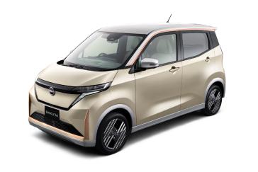 ベージュ系のボディカラーの「日産サクラ」を、無地の白いスタジオ背景の前で、車両の左前方から斜めに撮影した画像。フロントから側面にかけてのデザインが分かりやすく写っている。