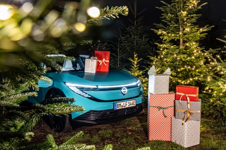 Blick durch Tannenzweige auf die Frontpartie eines türkisblauen Nissan LEAF, vor und auf dessen Fronthaube verpackte Weihnachtsgeschenke stehen
