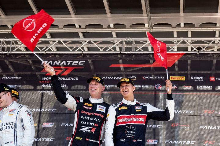 Two smiling racers in black and white Nissan NISMO race suits standing on a race stage waving red NISMO flags