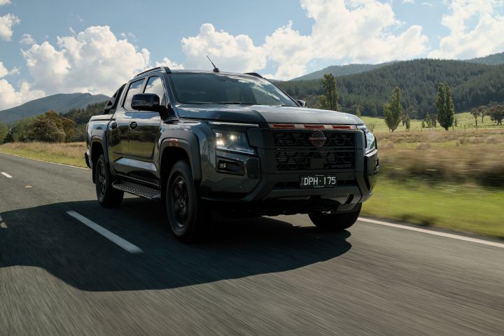 2026 Nissan Navara PRO-4X finished in Boulder Grey portraying a three-quarter cruising view beside open grassland with tub rails, side step and wheel-arch cladding.