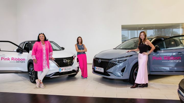 Die drei Frauen stehen verteilt um die zwei schräg zueinander ausgerichteten Autos, links steht ein weißer Nissan Juke, rechts ein grauer Nissan Qashqai. Auf beiden Fahrzeugen sind die Worte Pink Ribbon powered by Nissan zu lesen.