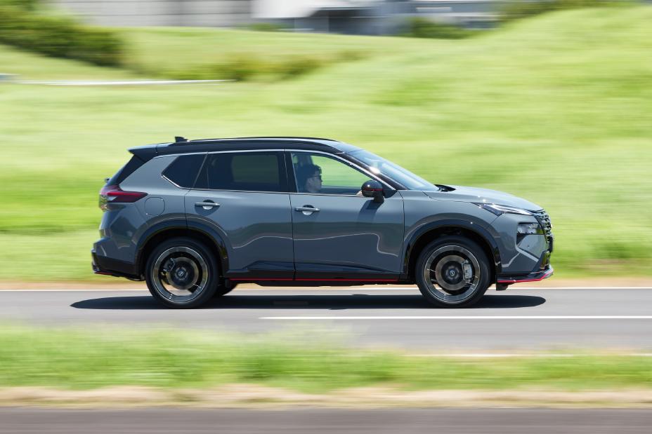 A grey Nissan X-Trail NISMO is driving on a road. The vehicle is captured from the side, with green grass and a blurred background indicating motion.