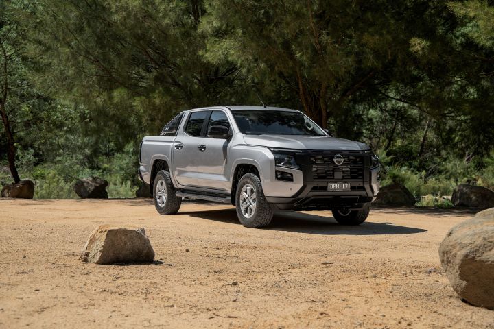 2026 Nissan Navara ST dual cab ute finished in Summit Silver captured from the front-left on sandy ground, with a foreground rock and tree-lined setting.