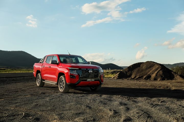 Wide front three-quarter view of a 2026 Nissan Navara ST‑X dual cab in Outback Red, stationary on gravel beside a low dirt mound under a blue sky with scattered clouds.