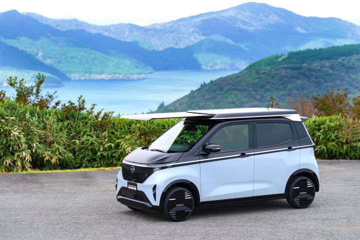 Against a backdrop of a lake and mountains, a light blue Nissan SAKURA is parked by the roadside, facing slightly right front. The solar roof extends forward prominently, with green shrubs around and white clouds scattered in the blue sky.