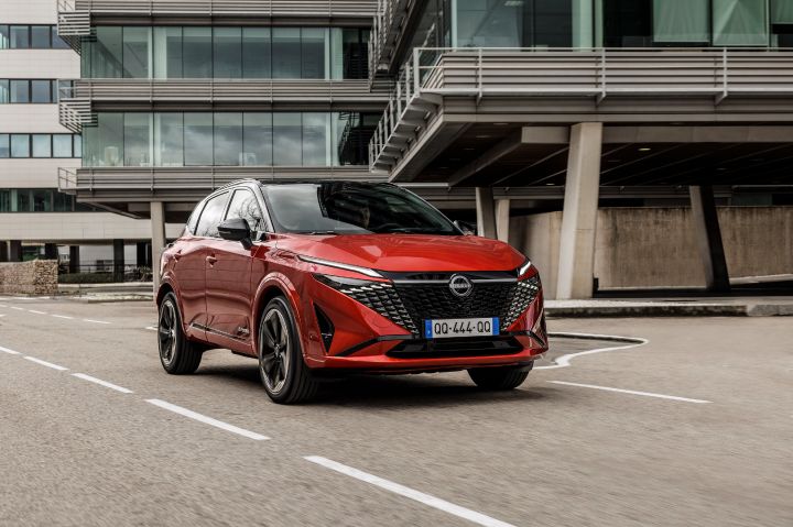 Een rode Nissan Qashqai met zwart contrasterend dak rijdt langs moderne kantoorgebouwen op straat, 3/4 vooraanzicht