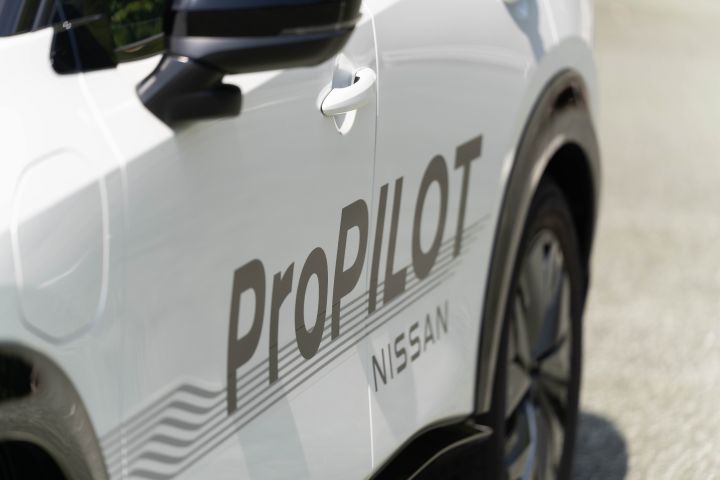 Seitenansicht eines weißen Nissan Ariya mit den Logos „ProPILOT” und „NISSAN” auf der Karosserie. Der Hintergrund ist unscharf und zeigt einen Teil einer Straße.