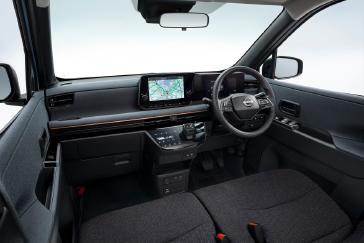 「日産サクラ」の車内を、前席の中央付近から斜め前に向けて撮影した画像。黒基調のインテリアとステアリング、センターディスプレイ、エアコン操作部が正面に見え、背景は白で統一されている。