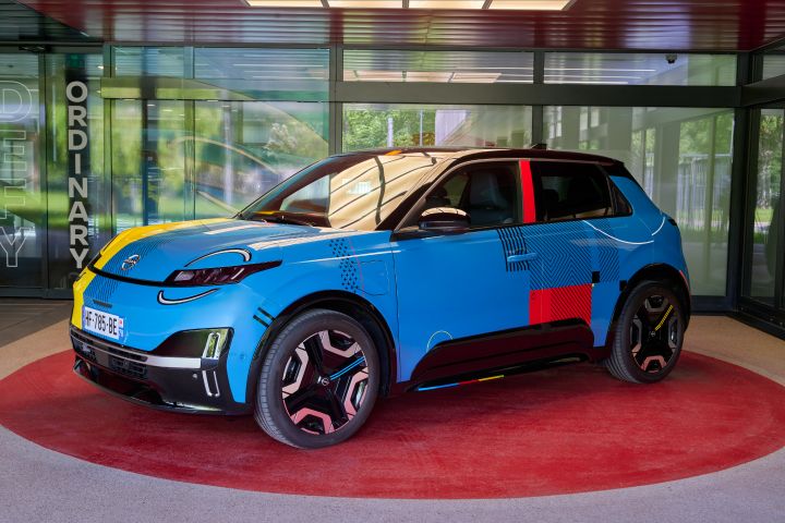 3/4 Frontansicht des blauen Nissan Micra Signature Edition Gewinnerfahrzeugs mit elektrischem Antrieb, schwarzem Dach, auffälligen roten, gelben und schwarzen grafischen Akzenten sowie sportlichen Felgen, ausgestellt in einem roten Kreis vor einer Glaswand.