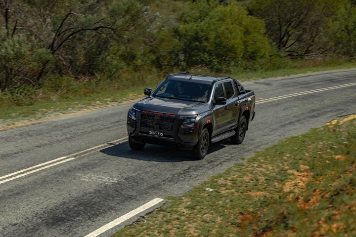 2026 Nissan Navara PRO-4X finished in Boulder Grey captured from a high front three‑quarter angle on country bitumen, approaching between double white lines with red grille accents, black wheels and side steps.