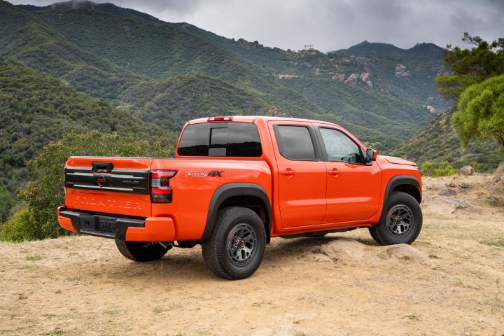 Right rear 3/4 angle of a red 2025 Nissan Frontier parked on a dirt cliff overlooking tree covered mountains.