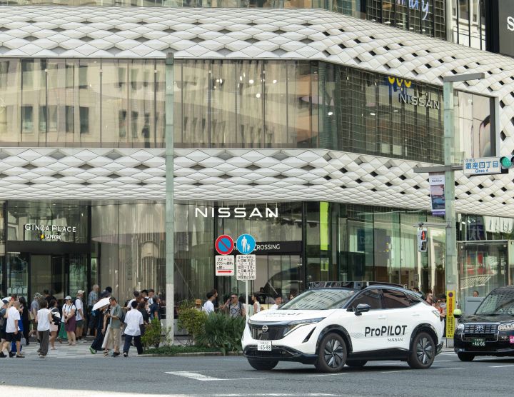 Ein weißer Nissan Ariya fährt vor dem NISSAN CROSSING-Gebäude, das eine Glasfassade mit markanten Mustern im oberen Bereich aufweist. Im Hintergrund sind weitere Gebäude und Fußgänger zu sehen.