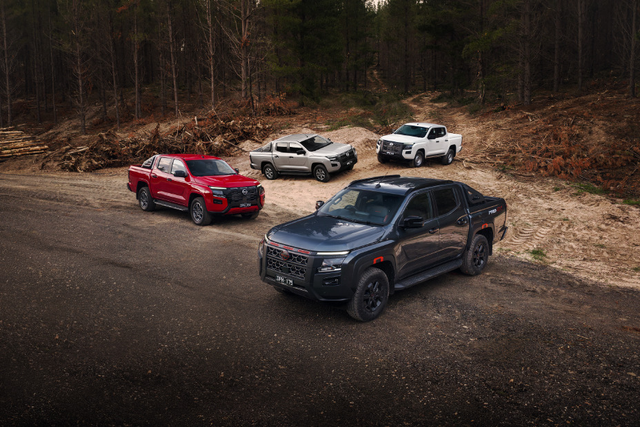 Four Nissan Navara variants arranged on a gravel roadside beside a forest clearing: a Navara PRO-4X in Boulder Grey at the front, a Navara ST-X in Outback Red to the left, a Navara ST in Summit Silver behind it, and a Navara SL in Alpine White at the rear right. Landscape