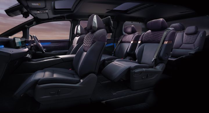 Interior view of the All-new ELGRAND, photographed from the front-left side looking toward the rear of the cabin. The premium purple interior features diamond-pattern seat upholstery arranged across three rows. The front area includes a large display integrated into the dashboard and a substantial center console, while the second row is equipped with individual captain’s chairs with the ottoman extended. Blue ambient lighting is visible along the door panels and cabin trim, and large glass roof panels span the ceiling above. A sunset sky can be seen through the windows, emphasizing the spacious and luxurious atmosphere of the interior.