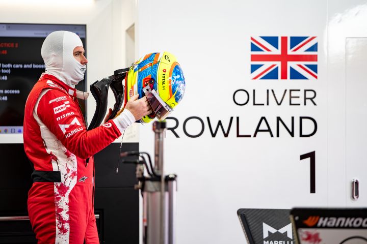 Oliver Rowland in seinem roten Rennanzug in der Garage des Nissan Formel E Teams, während er seinen fluoreszierenden gelb-blauen Helm aufsetzt.