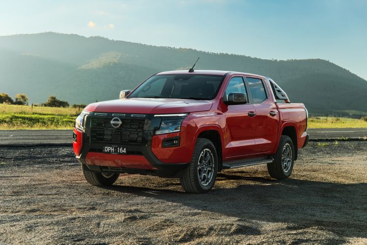 Front three-quarter view of a 2026 Nissan Navara ST-X dual cab in Outback Red, parked on a gravel turnout with mountain slopes and open countryside in the background.