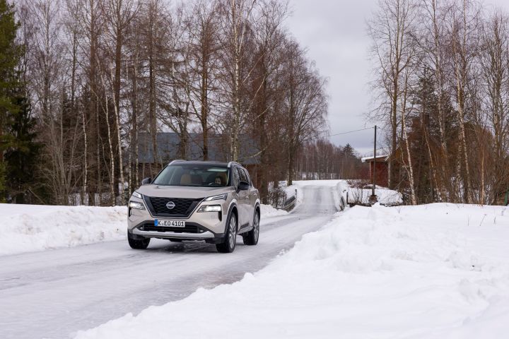 Silberner Nissan X‑Trail e‑Power e‑4ORCE fährt auf schneebedeckter Fahrbahn, im Hintergrund Bäume und Holzhäuser