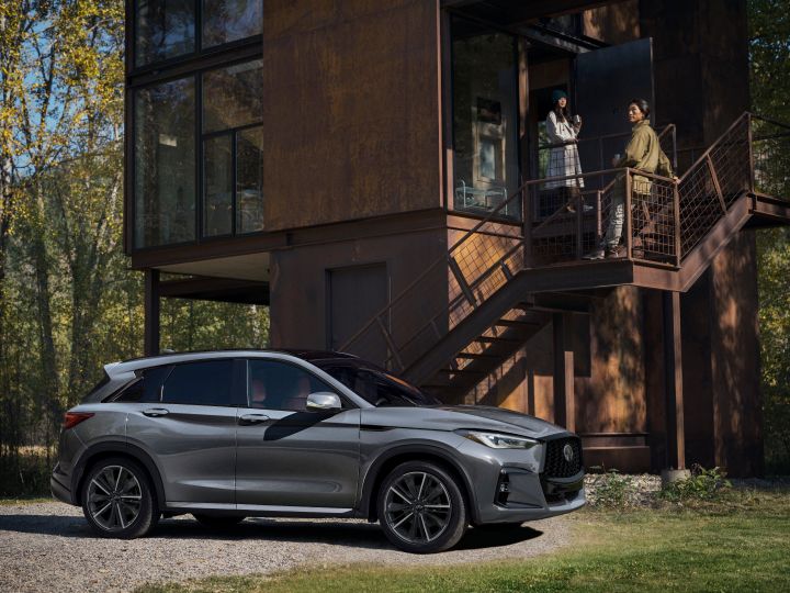 Right profile view of a 2025 INFINITI QX50 parked in front of a modern cabin, two women stand at the entrance of the cabin.