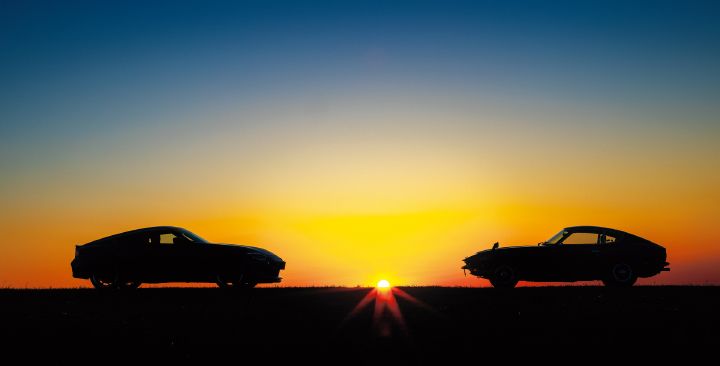 Silhouette image of a modern Unryu Green Fairlady Z and a classic S30 Fairlady Z at sunrise or sunset. The two vehicles are positioned on opposite sides of the horizon with the sun centered between them. The modern Fairlady Z on the left faces toward the right, while the first-generation S30 Fairlady Z on the right faces toward the left. Both vehicles appear as dark silhouettes, emphasizing their shared long-hood, short-deck proportions while highlighting the evolution of the Fairlady Z design across generations. A dramatic sky transitions from warm orange near the horizon to deep blue overhead, with the sun casting radiant beams that create a striking and symbolic scene celebrating the heritage of the Fairlady Z.