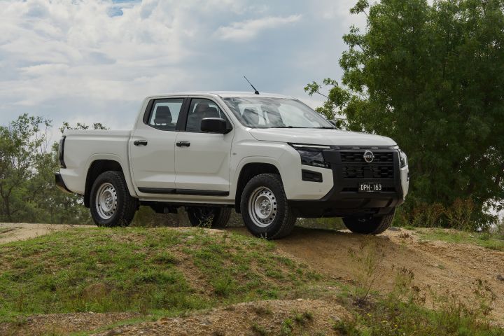 2026 Nissan Navara SL dual cab ute finished in Alpine White shown parked from the front-left on a grassy mound, featuring the side profile and black front-end treatment.