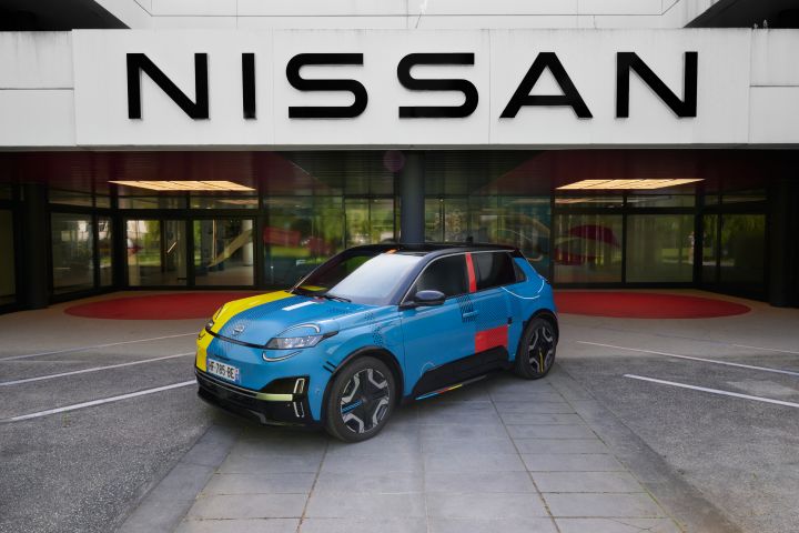 Nissan Micra azul com apontamentos em amarelo, vermelho e preto ao longo da viatura, visto de ¾ em frente a edifício a dizer “Nissan”