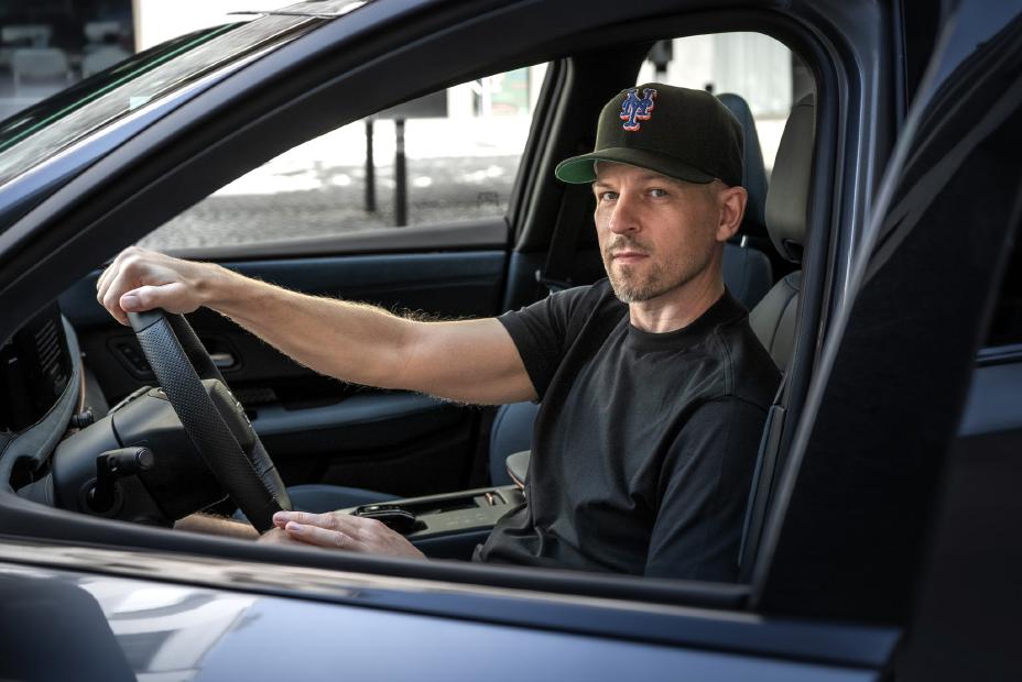 Le rappeur Stress est assis au volant d'une Nissan Ariya et regarde le spectateur à travers la fenêtre latérale ouverte.