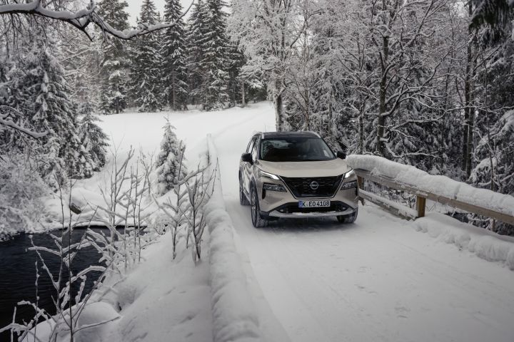 Silberner Nissan X‑Trail e‑Power e‑4ORcE fährt auf schneebedeckter Brücke mit verschneitem Wald im Hintergrund und einem Gewässer auf der linken Seite, Front