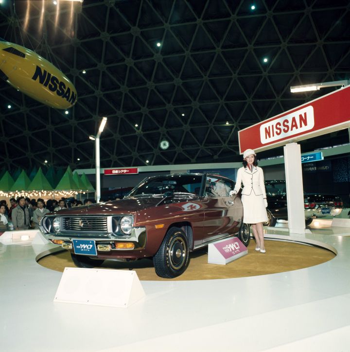 A vintage Nissan car displayed at an auto show. A person in a white outfit stands next to the vehicle on a circular platform.