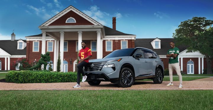 Two football players wearing college football jersey’s standing next to a gray Nissan Rogue in front of a large brick mansion.