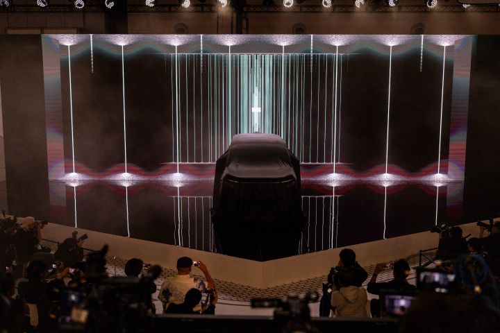 In a dark stage setting, the all-new Elgrand is positioned at the center, covered with a black cloth, while the background screen displays vertical light lines. In the foreground, members of the press are gathered, taking photos with cameras.