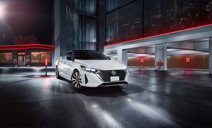 Auto Nissan blanco, estacionado de lado de noche, en un edificio de cemento con vidrio con luces rojas