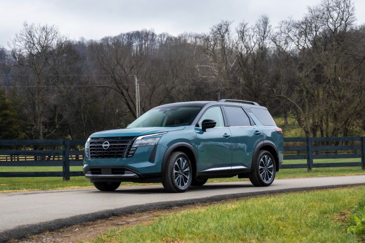 A teal 2026 Nissan Pathfinder parked on a concrete road in front of black wooden fence with trees in the background and grey skies