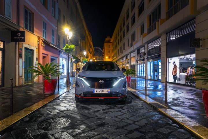 Vue frontale d'une Nissan Ariya NISMO gris-bleu avec des accents de design rouge, roulant de nuit le long d'une rue commerçante étroite et éclairée.