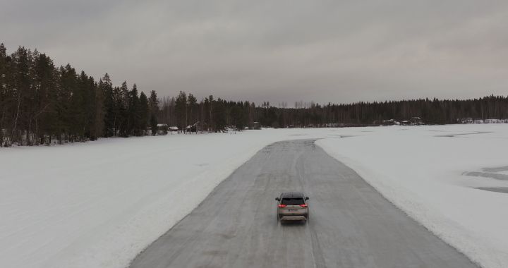 Silberner Nissan X‑Trail e‑Power e‑4ORcE fährt auf schneebedecktem Eissee mit verschneiten Bäumen und Holzhütten im Hintergrund, Heckansicht, kleines Auto in weißer Landschaft, Drohnen-Perspektive