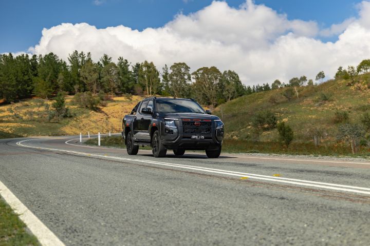 2026 Nissan Navara PRO-4X finished in Boulder Grey driving a sweeping highway section with rolling hills, guide posts and bright cloud.