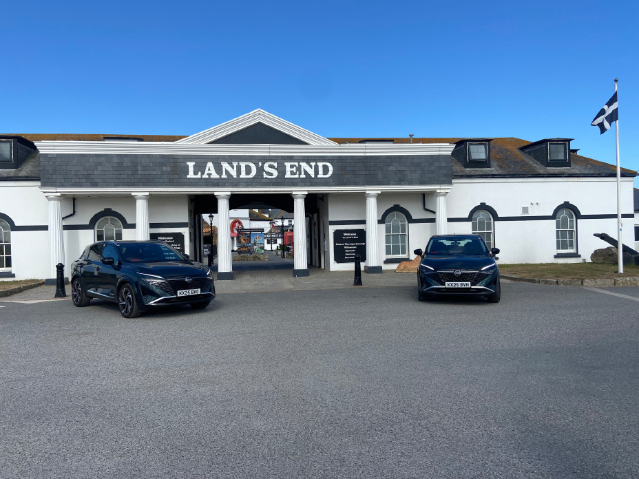 Zwei dunkelgrüne Nissan Qashqai mit e‑POWER Antrieb der dritten Generation stehen an einem weißen Gebäude mit dem großen Schriftzug Lands End, Start der 874-Meilen-Reise nach John o'Groats.