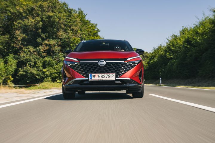 Frontalaufnahme eines fahrenden Nissan Qashqai in Fuji Sunset Red vor blauem Himmel und grünen Bäumen