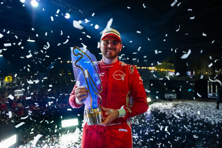 O piloto Oliver Rowland vestido com o uniforme vermelho da Nissan e um boné branco segura um troféu dourado e prateado