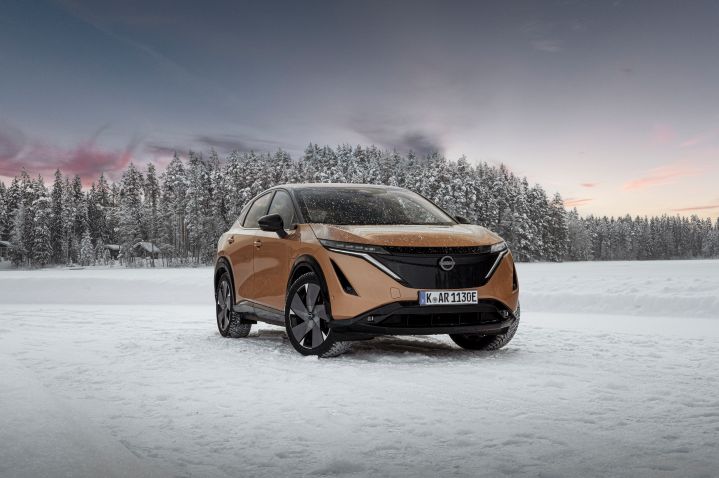Elektroauto Nissan Ariya e‑4ORCE in Akatsuki Copper mit Dach und Außenspiegel in Pearl Black auf Eissee mit schneebedeckten Bäumen im Hintergrund, 3/4 Front statisch