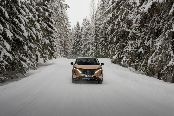 Elektroauto Nissan Ariya e‑4ORCE in Akatsuki Copper mit Dach und Außenspiegel in Pearl Black fährt auf schneebedeckter Straße mit verschneiten Bäumen auf beiden Seiten, Front