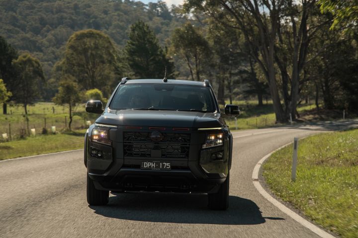 2026 Nissan Navara PRO-4X finished in Boulder Grey pictured rounding a gentle left-hand bend with open meadow, white guide post and scattered trees in afternoon light.