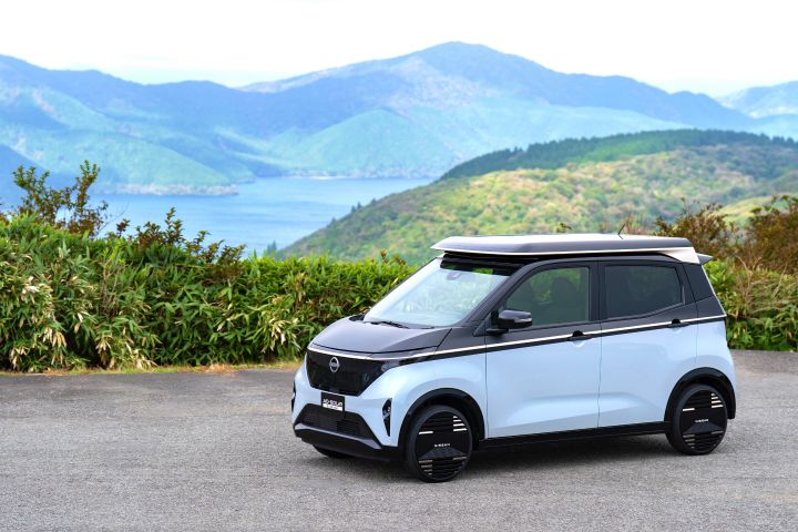 Against a backdrop of a lake and mountains, a light blue Nissan SAKURA is parked by the roadside, facing slightly right front. Green shrubs surround the area, and the sky is blue with scattered white clouds.