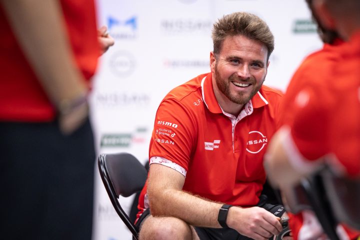 Oliver Rowland sitzt in seinem roten Nissan Formula E Team-Poloshirt lächelnd in der Teamgarage.