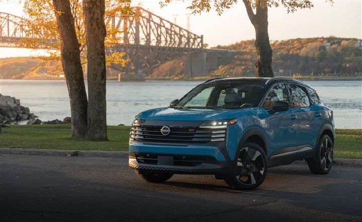 Front-left view of a blue 2025 Nissan Kicks parked near water at sunset, with a bridge and tree-covered hills in the background.