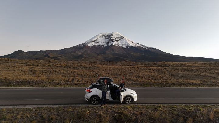 Guilherme e Camila estão ao lado de um carro branco, com a porta aberta, estacionado à beira da estrada. Ambos levantam os braços, comemorando. Ao fundo, o vulcão Chimborazo domina a paisagem, com seu cume coberto de neve contrastando com o terreno árido ao redor. O céu está limpo