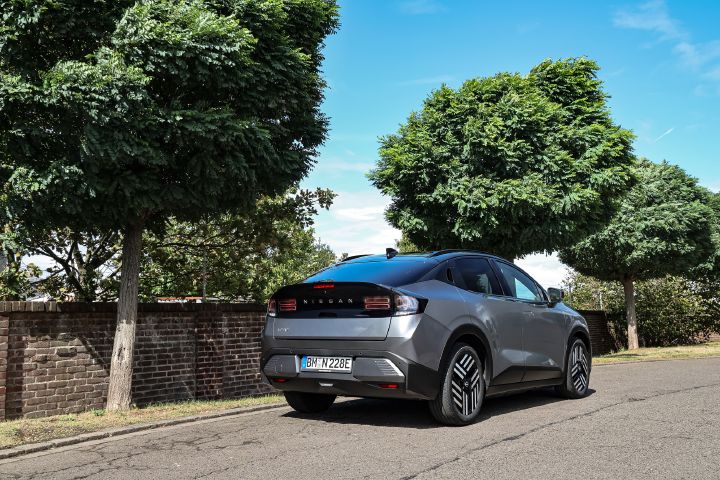 3/4 Heckansicht eines grauen Nissan Leaf, der vor Bäumen und einer halbhohen Mauer steht